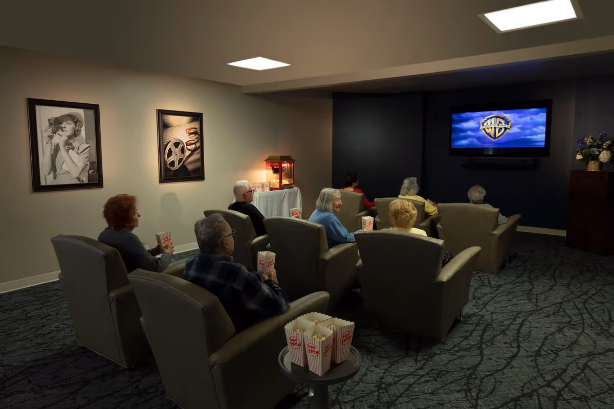 A group of elderly people seated in comfortable armchairs in a dimly lit room watching a movie on a wall-mounted TV. Each person has a container of popcorn. The room has framed black and white photos on one wall, a popcorn machine on a table, and a floral arrangement on a cabinet near the TV.