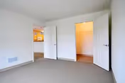 Empty room with beige carpet and white walls, featuring two open doors; one leading to a walk-in closet with shelves and the other opening into a hallway with a view of a kitchen area.