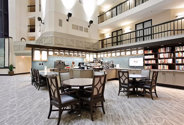A spacious and well-lit common area with round tables and chairs arranged for seating. The area features a counter with coffee machines and other equipment, and a wall lined with bookshelves filled with books. The space has high ceilings and multiple levels with railings visible above.