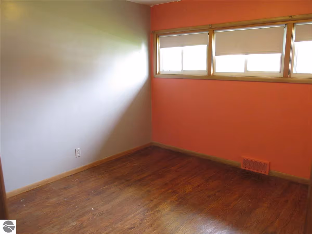 Empty small room with hardwood floor, a coral accent wall and three high windows with shades.