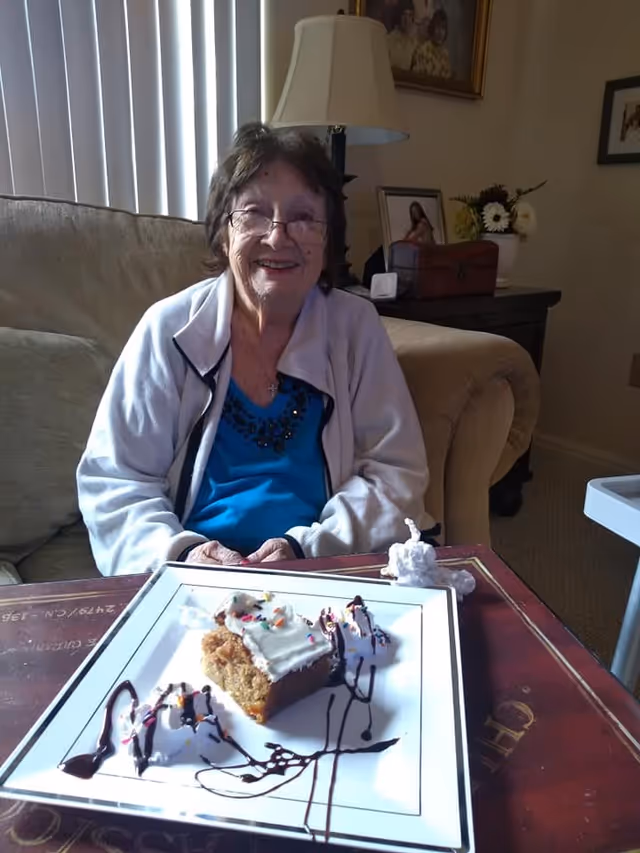 An elderly woman wearing glasses and a light jacket sits on a beige couch in a cozy living room. In front of her is a table with a plate holding a piece of cake topped with white frosting and colorful sprinkles, accompanied by whipped cream drizzled with chocolate syrup. Behind her, there is a lamp, framed pictures, and a flower arrangement on a side table.