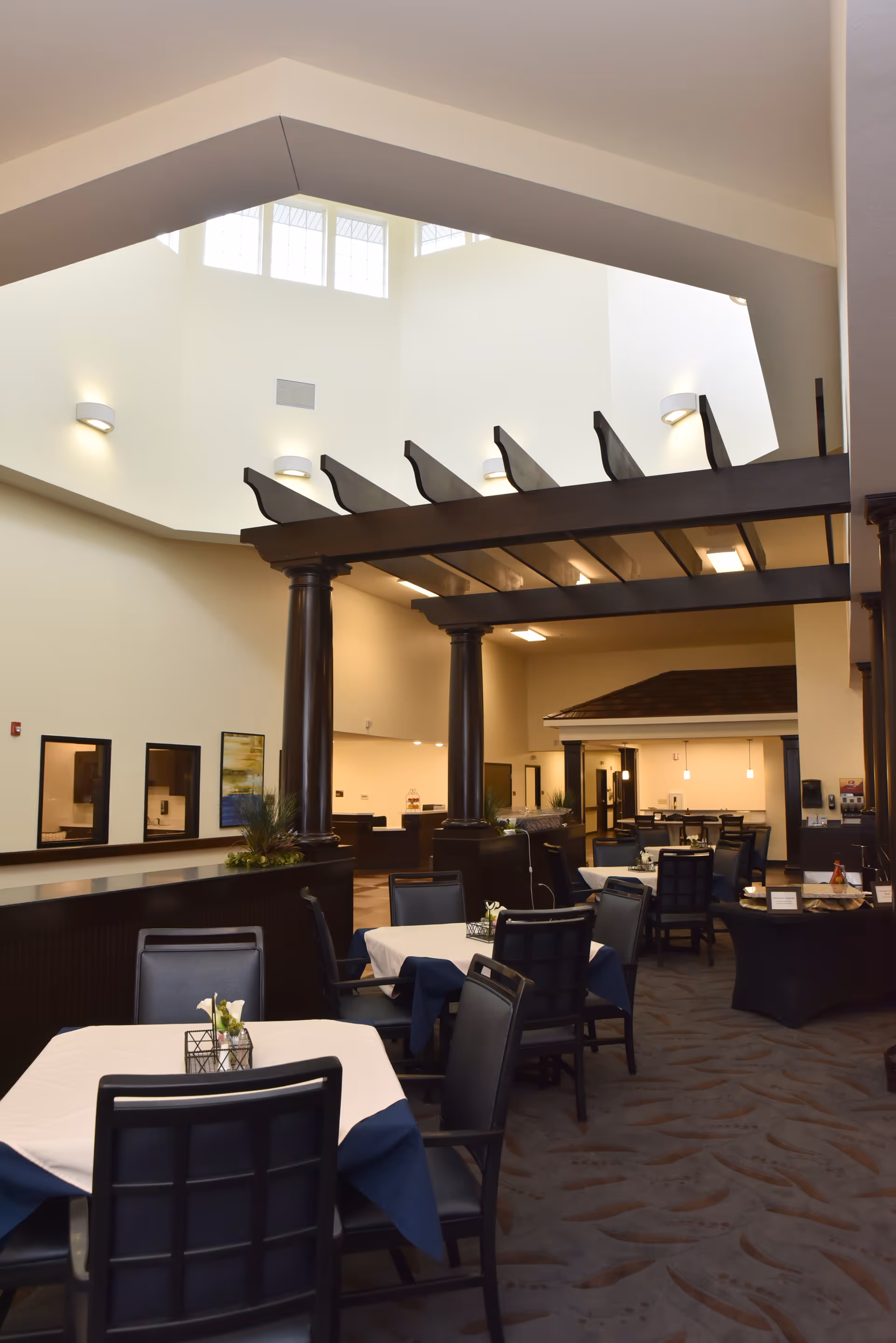 Interior view of a dining area in Orchard Pointe Health Campus featuring tables with white and blue tablecloths, black chairs, dark wooden columns, and a high ceiling with windows allowing natural light.