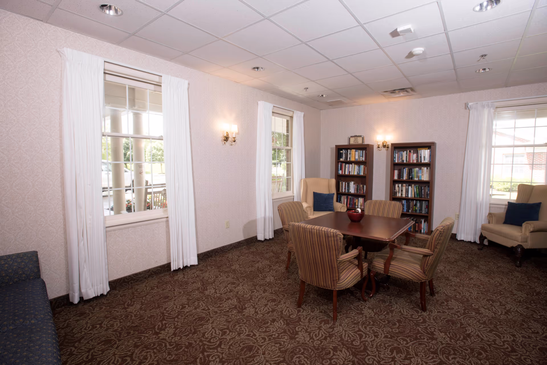A cozy interior room with patterned carpet and light-colored wallpaper. The room features a square wooden table surrounded by four striped upholstered chairs. There are two beige armchairs with dark blue cushions near the windows, and two bookshelves filled with books against the far wall. White curtains frame the windows, and wall sconces provide warm lighting.