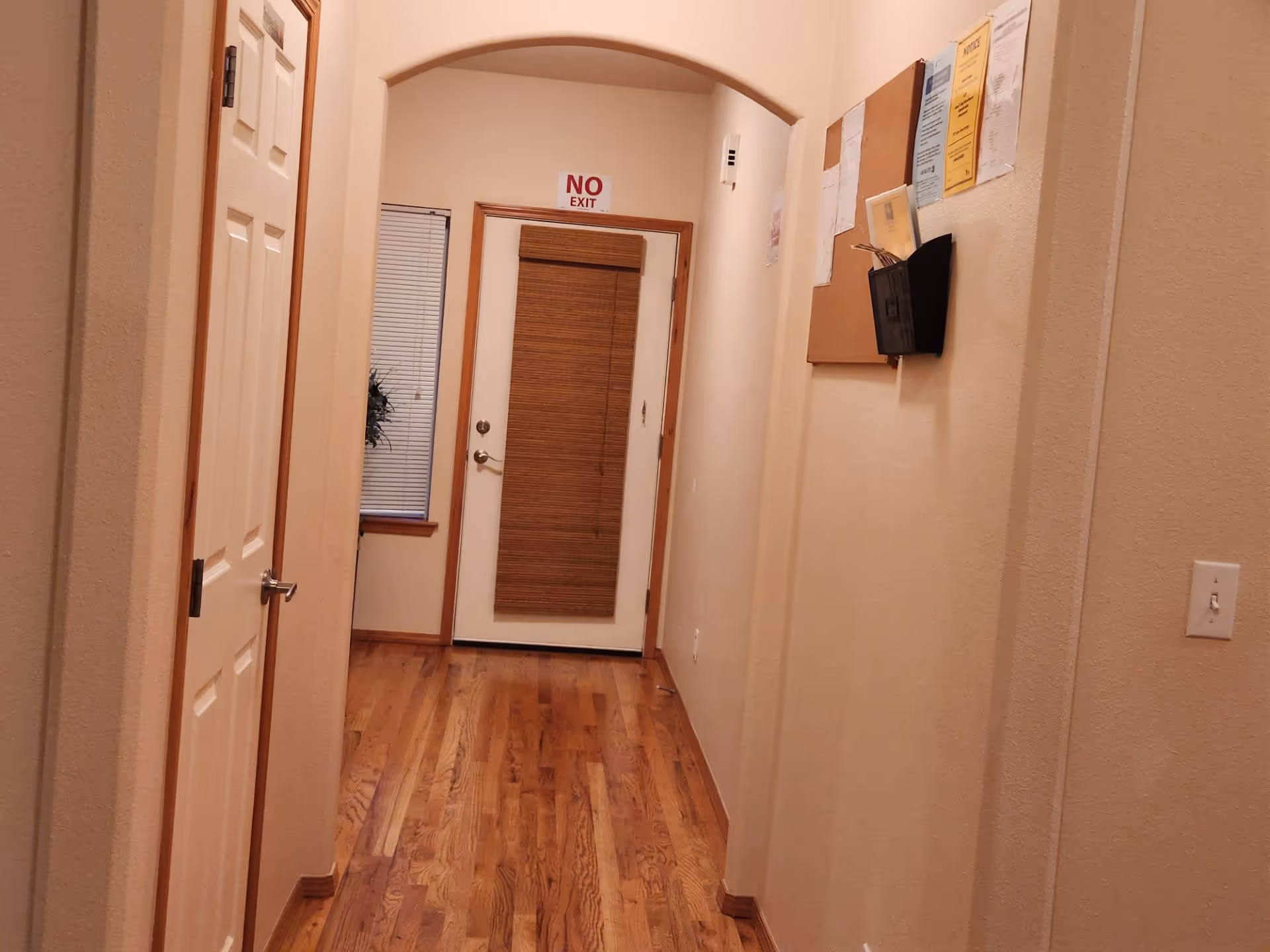 A hallway with wooden flooring and beige walls. On the left side, there is a closed white door. At the end of the hallway, there is a door with a wooden blind and a window with blinds to its left. Above the door, a sign reads 'NO EXIT'. On the right wall, there is a bulletin board with various papers pinned to it and a black wall-mounted holder. A light switch is visible on the right wall near the foreground.