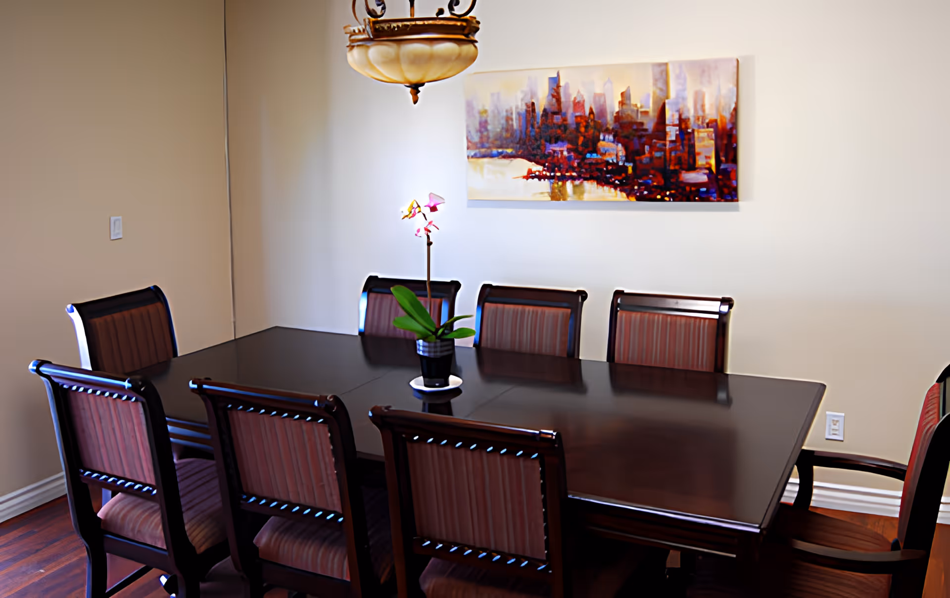 A dining room with a dark wooden rectangular table surrounded by eight matching chairs with striped upholstery. A small potted orchid is placed in the center of the table. A colorful cityscape painting hangs on the beige wall behind the table, and a decorative ceiling light fixture is visible above.