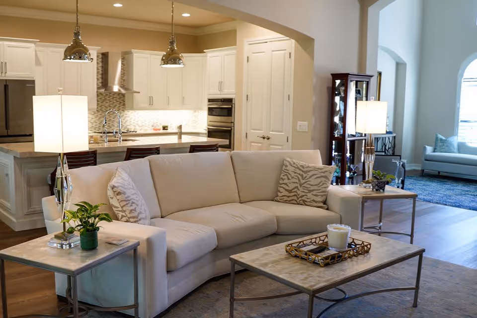 A modern living room with a beige sectional sofa adorned with patterned pillows. In front of the sofa is a rectangular coffee table with a candle and a decorative tray. Two side tables with lamps and small plants flank the sofa. The background shows a kitchen with white cabinetry, a tiled backsplash, stainless steel appliances, and pendant lights hanging over a breakfast bar with stools. The room has wooden flooring and an open layout leading to another seating area with a blue rug and a large window.