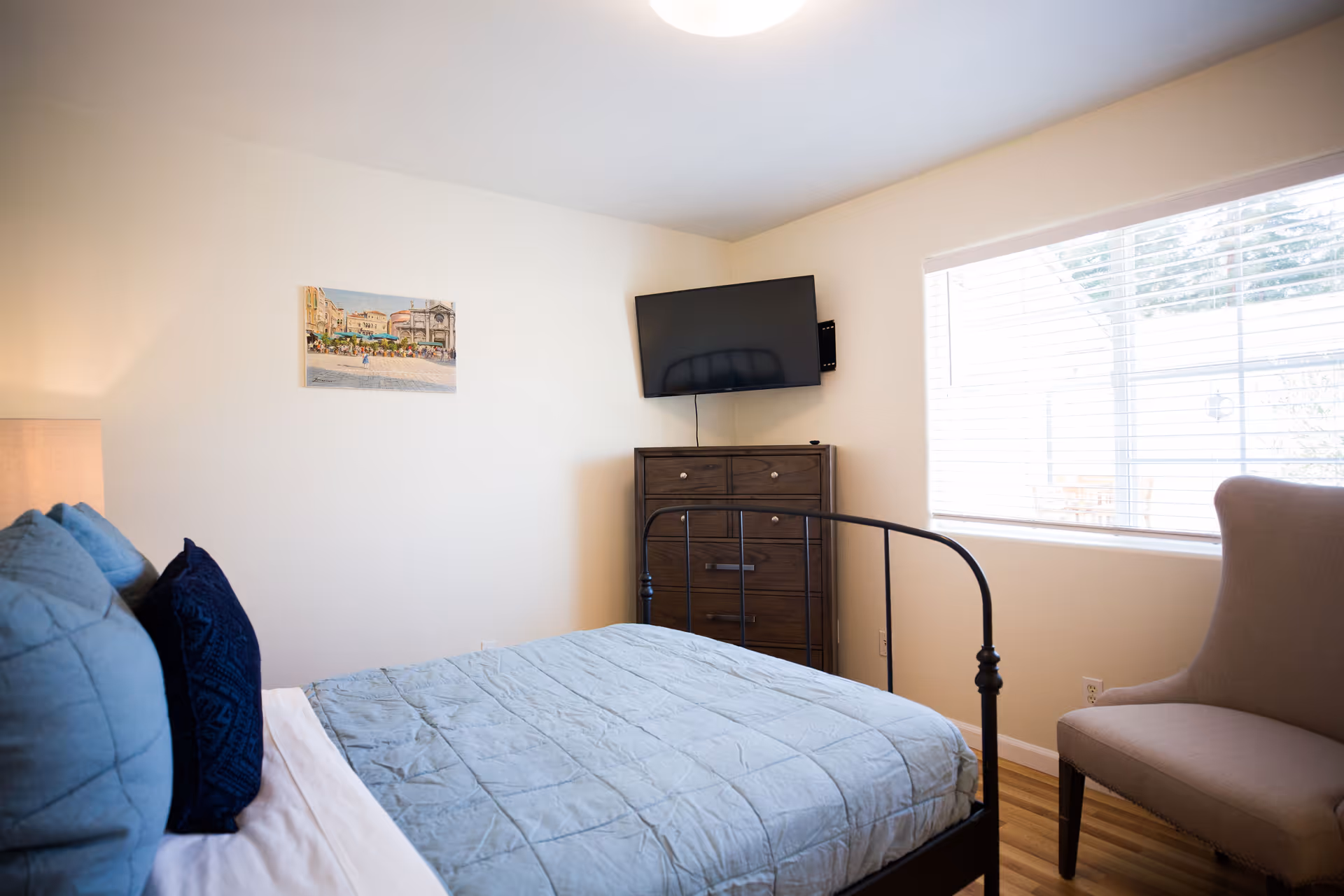 A bedroom with a bed covered in a light blue quilt and blue pillows, a wooden dresser with a flat-screen TV mounted above it, a beige armchair near a window with white blinds, and a framed picture on the wall.