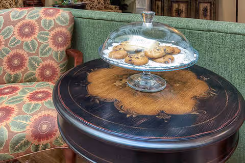 A close-up of a round wooden table with intricate designs, holding a glass cake stand with a dome lid containing several chocolate chip cookies. Next to the table is a cushioned chair with a floral patterned fabric in shades of green, red, and beige.