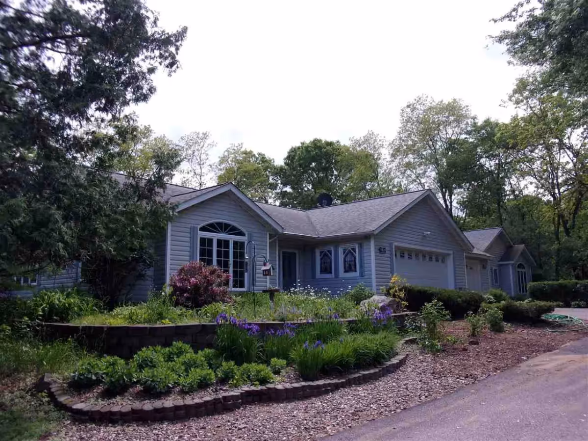 A single-story residential building with light gray siding and a dark gray roof, surrounded by lush greenery and a well-maintained garden with various plants and flowers. The building has multiple windows, a garage, and a driveway leading up to it, set against a backdrop of tall trees under a bright sky.