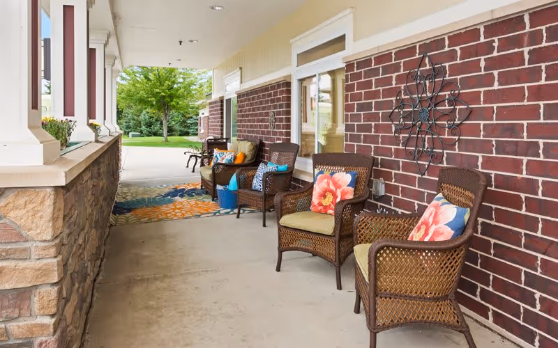 Covered outdoor patio area with wicker chairs featuring colorful floral and patterned cushions, a decorative metal wall art on a brick wall, and a view of green trees and grass in the background.