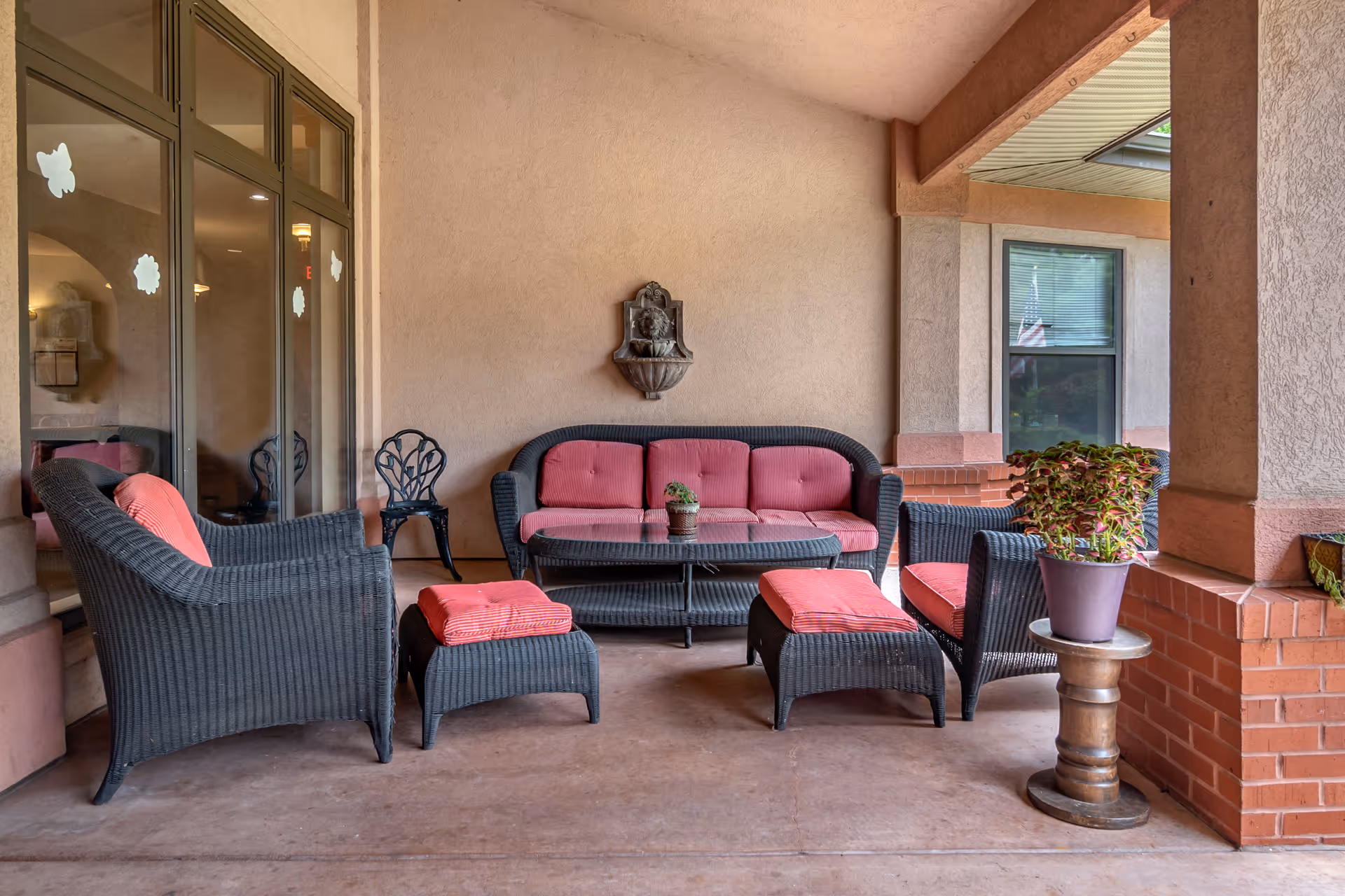 Covered outdoor patio area with black wicker furniture including a sofa, two chairs, two ottomans, and a coffee table, all with red cushions. There is a small potted plant on the coffee table and another potted plant on a small round side table. The patio has beige walls, brick pillars, and large windows with decorative decals.
