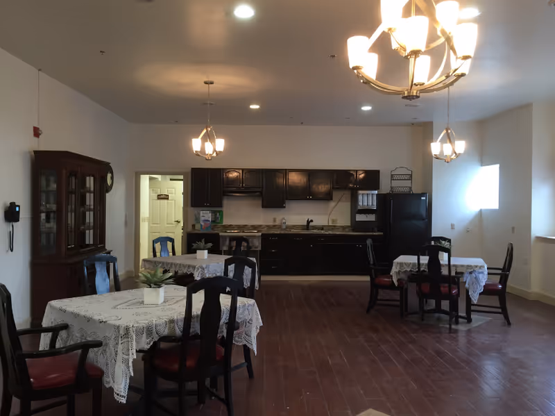 Interior view of a communal dining area with three round tables covered with white lace tablecloths and small potted plants as centerpieces. Each table is surrounded by dark wooden chairs with red cushions. The back wall features a kitchen area with dark cabinets, a countertop, a sink, and a black refrigerator. Three ceiling light fixtures with multiple bulbs illuminate the room.