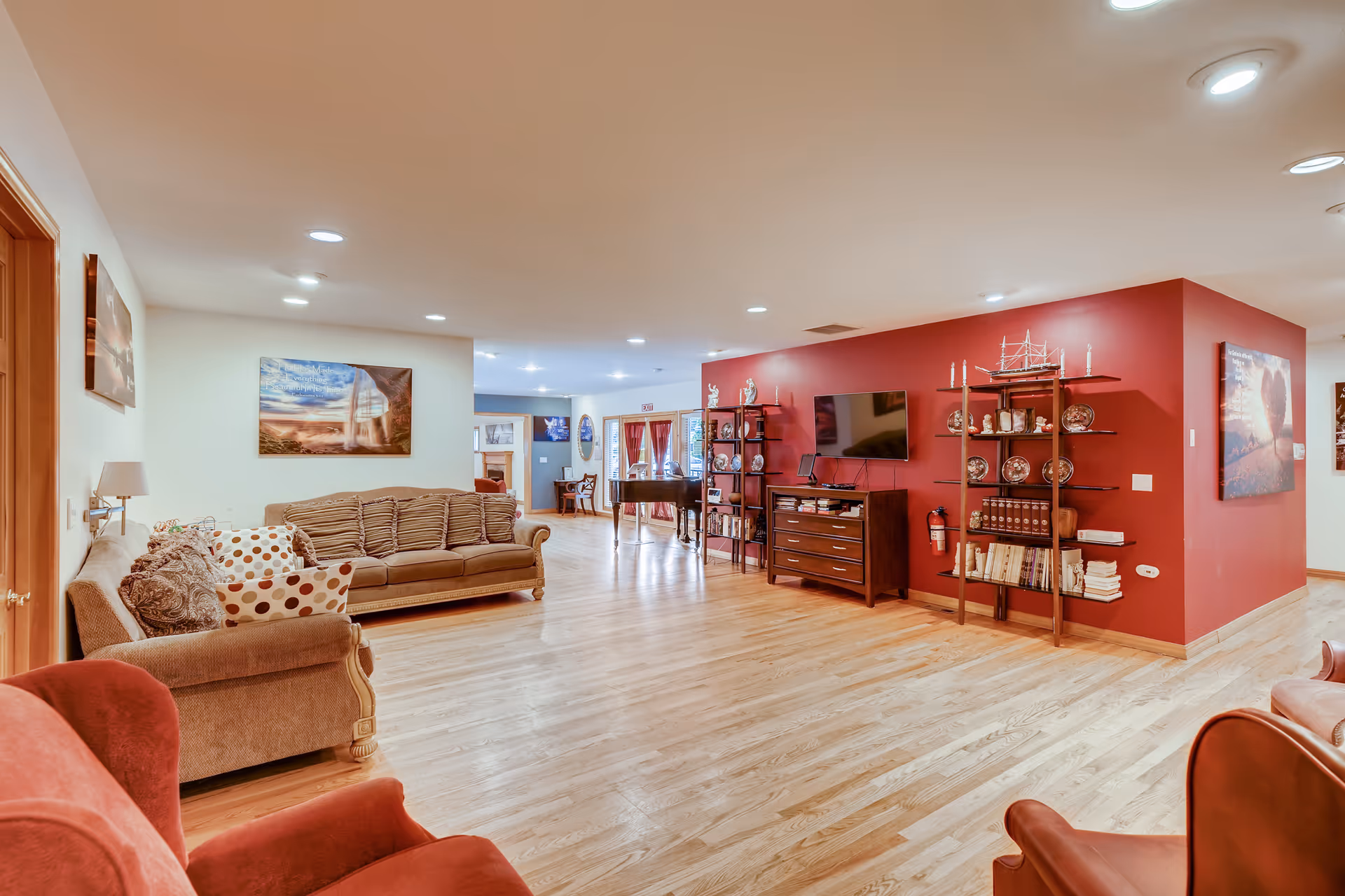 A spacious living room area with wooden flooring, beige and red upholstered sofas, a red accent wall with shelves displaying decorative plates and figurines, a flat-screen TV mounted on the wall, and framed artwork on the walls. The room is well-lit with recessed ceiling lights and has a cozy, inviting atmosphere.