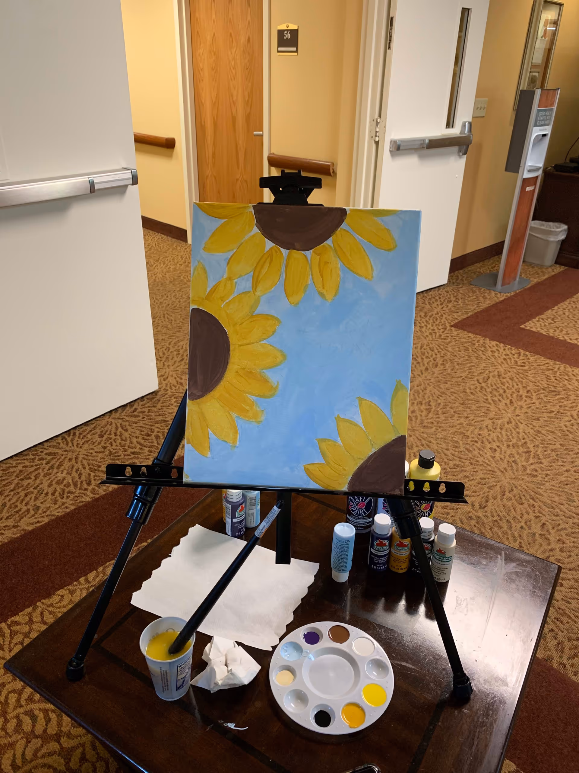 An easel on a wooden table in a hallway with a painting of three large sunflowers with yellow petals and brown centers against a blue background. The table holds a paint palette with various colors, paint bottles, a cup with a paintbrush, and paper towels. The hallway has beige walls, carpeted floor, and several doors.