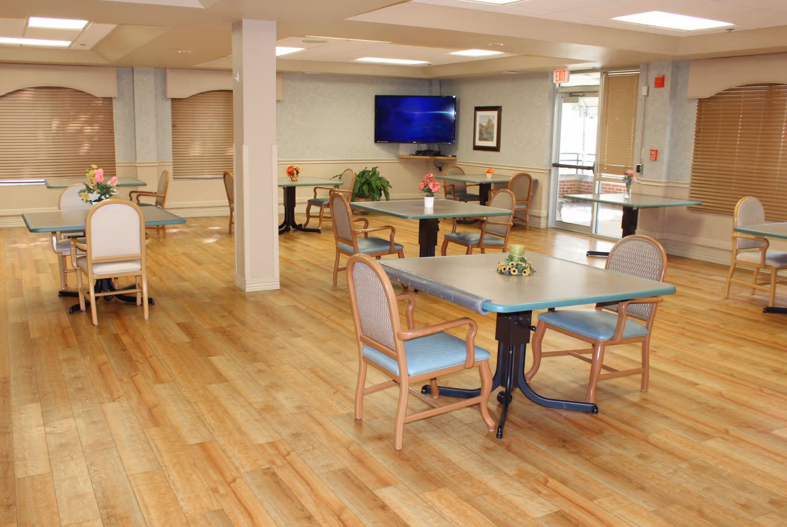 Communal dining area with multiple tables and chairs on wood-look flooring, floral centerpieces, and a wall-mounted television.