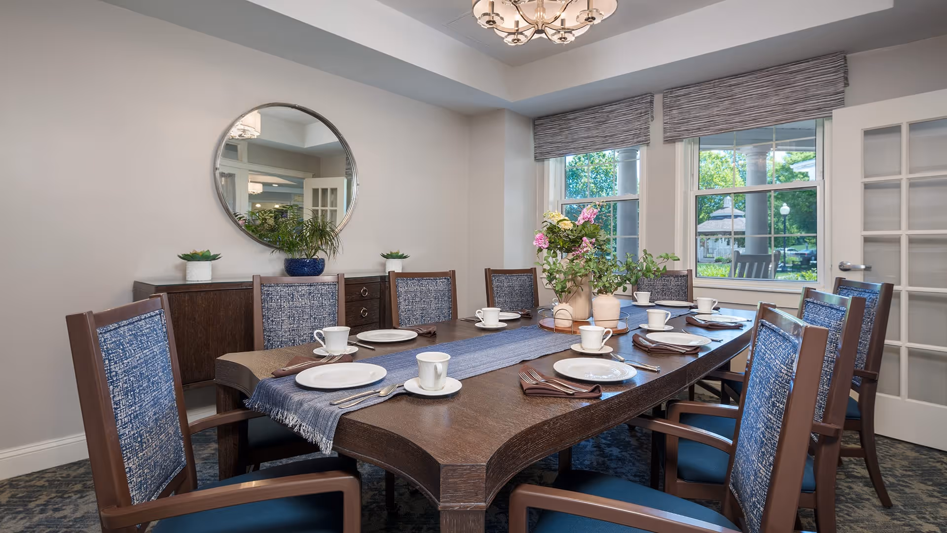 A well-lit dining room with a large wooden table set for eight people. The table has white plates, cups, saucers, and brown napkins arranged neatly. A blue table runner and a floral centerpiece with pink and white flowers add decoration. The room features a round mirror on the wall, a sideboard with plants, large windows with gray blinds, and a chandelier hanging from the ceiling.
