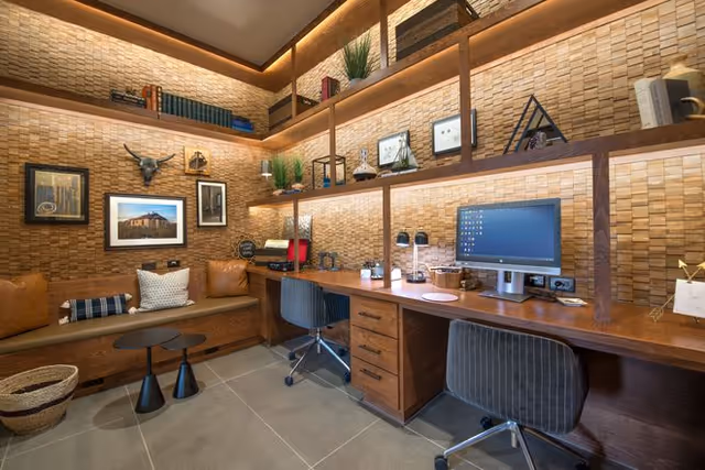 A cozy office or study room with a long wooden desk featuring two office chairs, a computer monitor, and various desk accessories. The walls are decorated with textured wood panels and floating shelves holding books, plants, and decorative items. There is a built-in bench with cushions along one wall, accompanied by two small round tables and a woven basket on the floor.