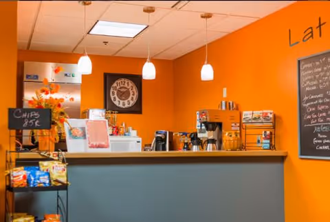Interior view of a small snack and coffee counter area with an orange wall background. The counter has a coffee machine, a glass container with snacks, and a menu board on the right side. There is a rack with bags of chips priced at 85 cents on the left side. Three pendant lights hang from the ceiling above the counter.