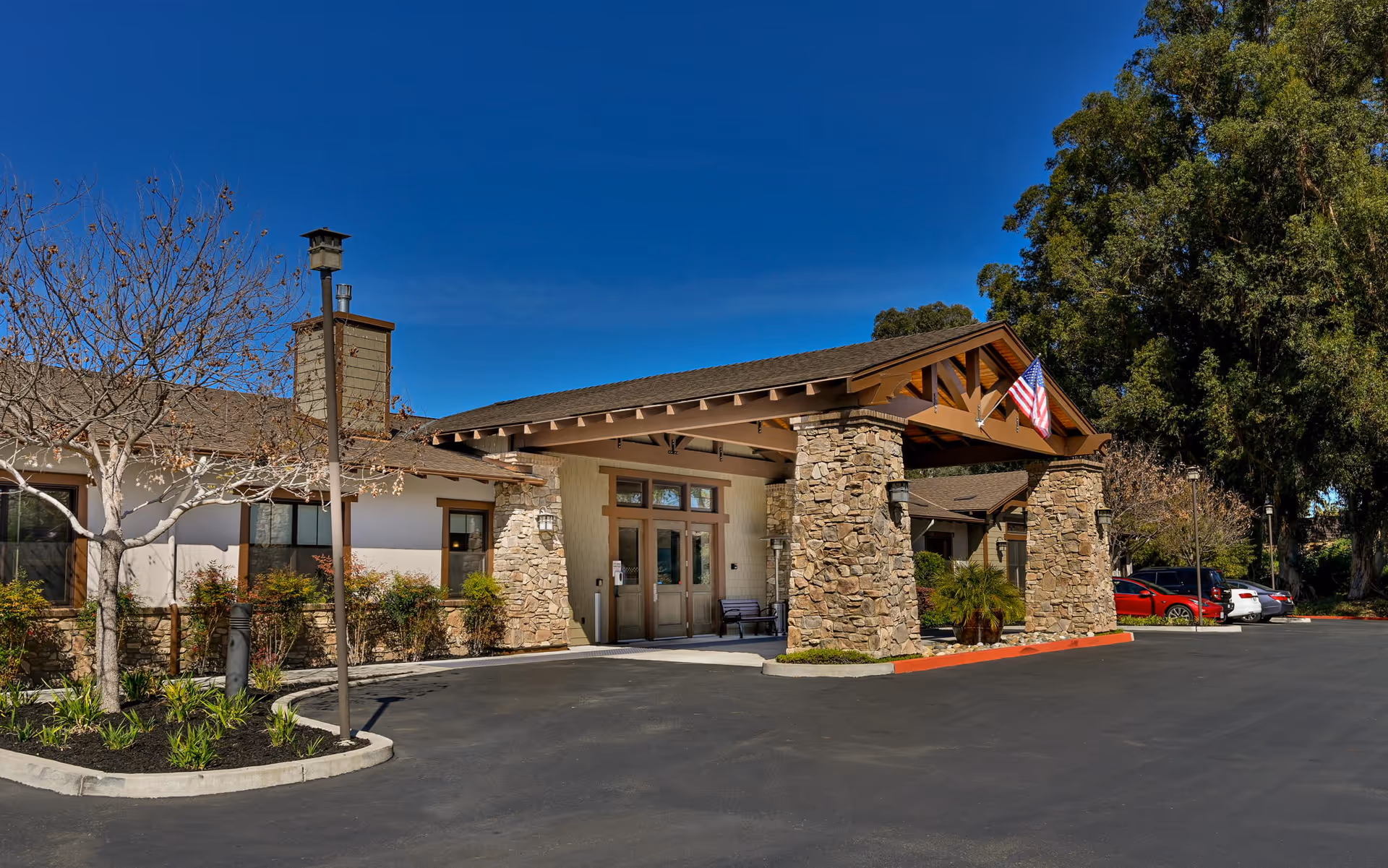 Front entrance of a memory care building with stone pillars, a covered entryway, American flag, landscaping, and a parking area.