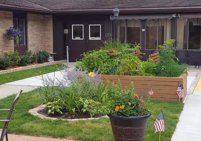 Outdoor garden area at St Michael's Assisted Living featuring raised wooden planter boxes and a circular flower bed with various plants and flowers. Two small American flags are placed in the garden and a large potted plant. The building entrance with dark double doors labeled 'Main Entrance' is visible in the background along with windows and a hanging flower basket.