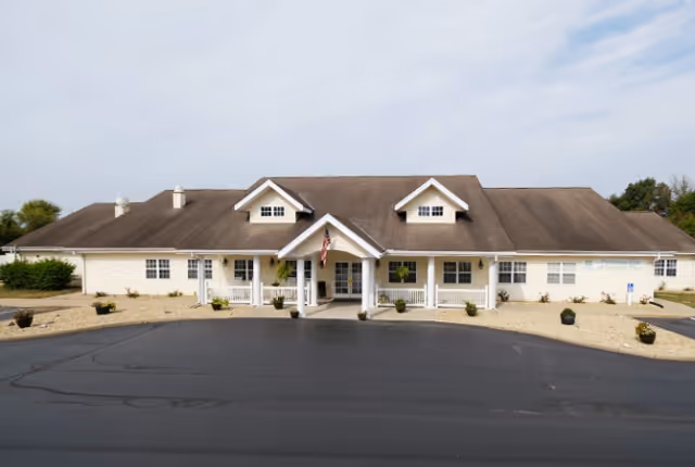 Front exterior view of a single-story building with a brown roof and white siding, featuring a covered entrance with an American flag and several windows. The surrounding area has gravel landscaping with small plants and a paved driveway in front.