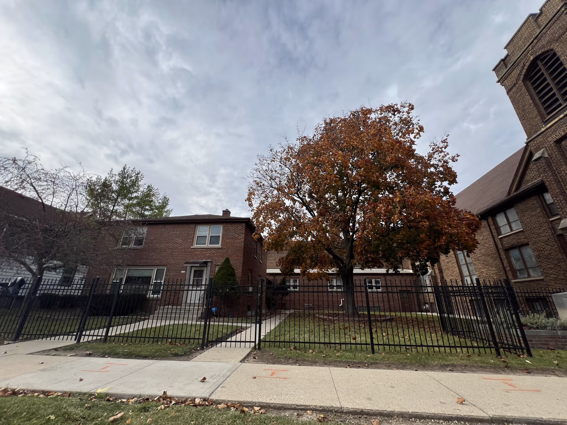 Exterior view of a brick residential building with a fenced front yard and a large tree with autumn-colored leaves. The sky is cloudy and there is a sidewalk in front of the property.