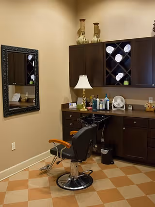 Interior view of a salon or grooming area with a black salon chair in front of a counter with various hair care products, a lamp, and a small fan. Above the counter are dark wooden cabinets with rolled white towels stored in a wine rack style section. A large decorative mirror is mounted on the adjacent wall. The floor has a checkered pattern in beige and light brown tones.