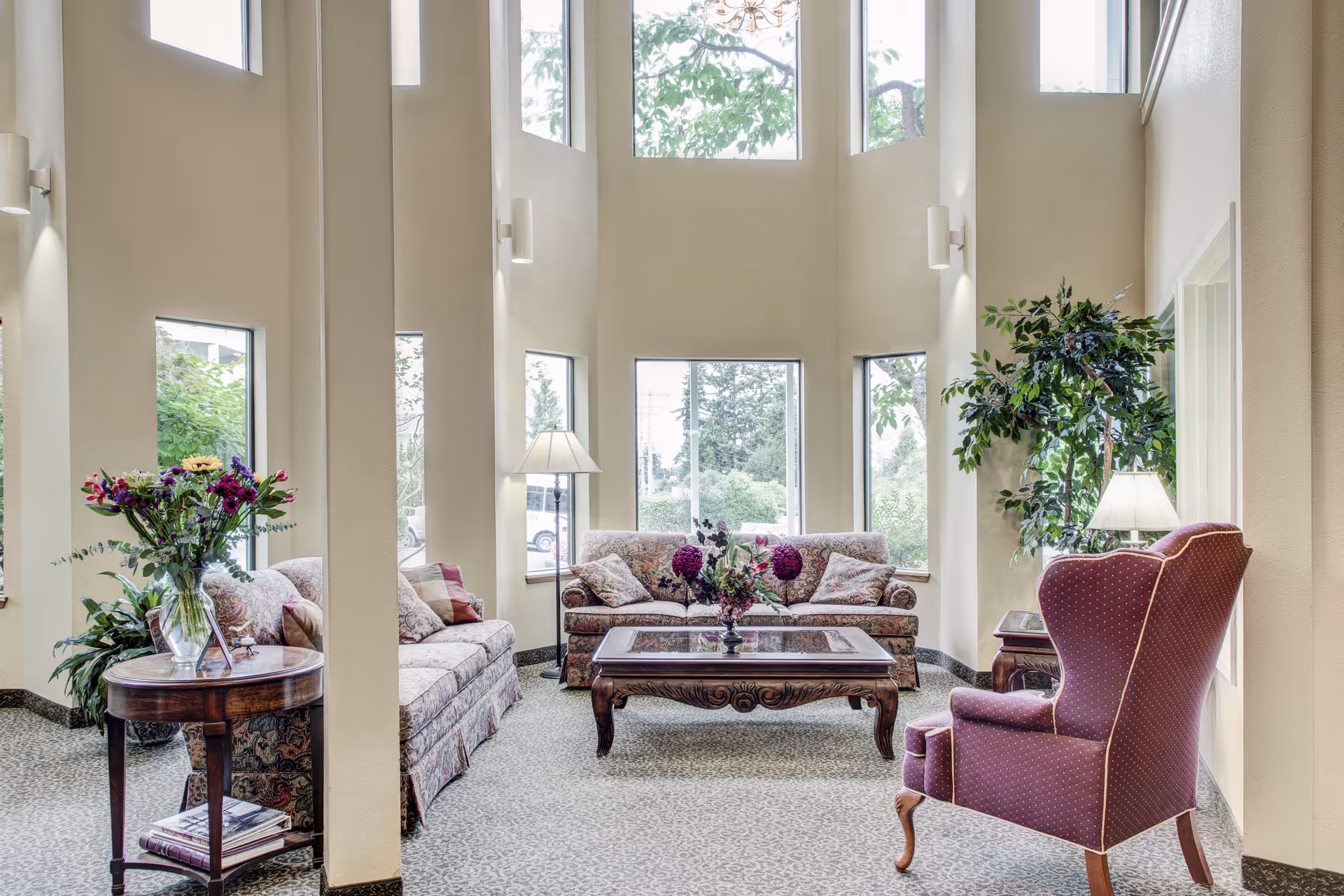 A bright and spacious living room area with high ceilings and multiple tall windows letting in natural light. The room features two patterned sofas with cushions, a wooden coffee table with a floral centerpiece, a purple upholstered armchair, a side table with a lamp, and a round wooden table with a vase of flowers. There are also green plants placed around the room.