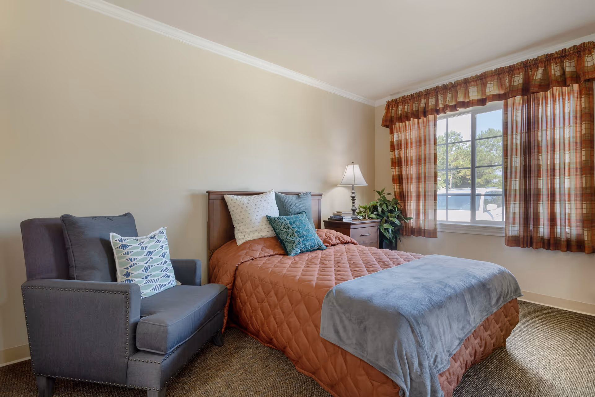 A cozy bedroom with a single bed covered in a rust-colored quilt and a gray blanket at the foot. The bed has three pillows, two white and one teal. Next to the bed is a wooden nightstand with a lamp, some books, and a green plant. To the left of the bed is a gray armchair with a decorative pillow featuring a fish pattern. A window with plaid curtains lets in natural light, showing trees and a parked vehicle outside.