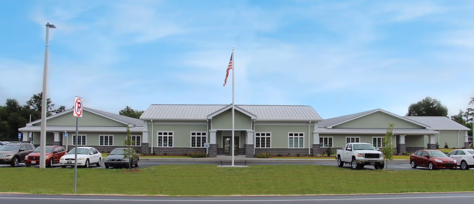 Front exterior view of a single-story building with a light green facade, white trim, and a metal roof. There is a flagpole with an American flag in front of the building, a parking lot with several cars, and a no parking sign near the street. The sky is clear with some clouds.