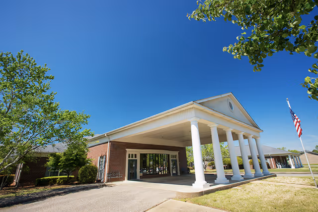 Exterior view of a single-story brick building with a large covered entrance supported by white columns, surrounded by trees and a clear blue sky. An American flag is visible on a flagpole to the right.