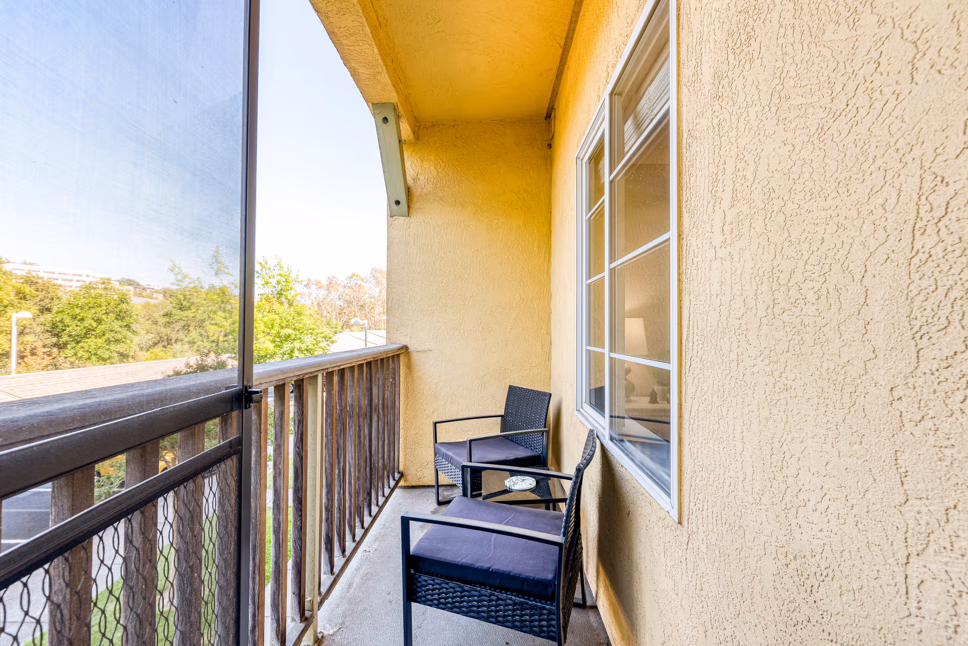 A narrow outdoor balcony with two black wicker chairs with cushions and a small glass-top table between them. The balcony has a wooden railing with a mesh screen on the left side and a textured yellow wall on the right with a window. Trees and a clear sky are visible in the background.