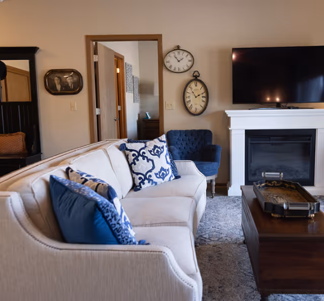 Cozy living room with a beige sofa topped with blue patterned pillows, a blue armchair, wall clocks, a TV over a white fireplace, and a wooden coffee table.