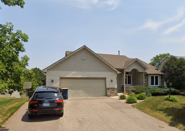 Front exterior view of a single-story residential building with a two-car garage, a driveway with a black SUV parked, and a well-maintained lawn with shrubs and trees under a partly cloudy sky.