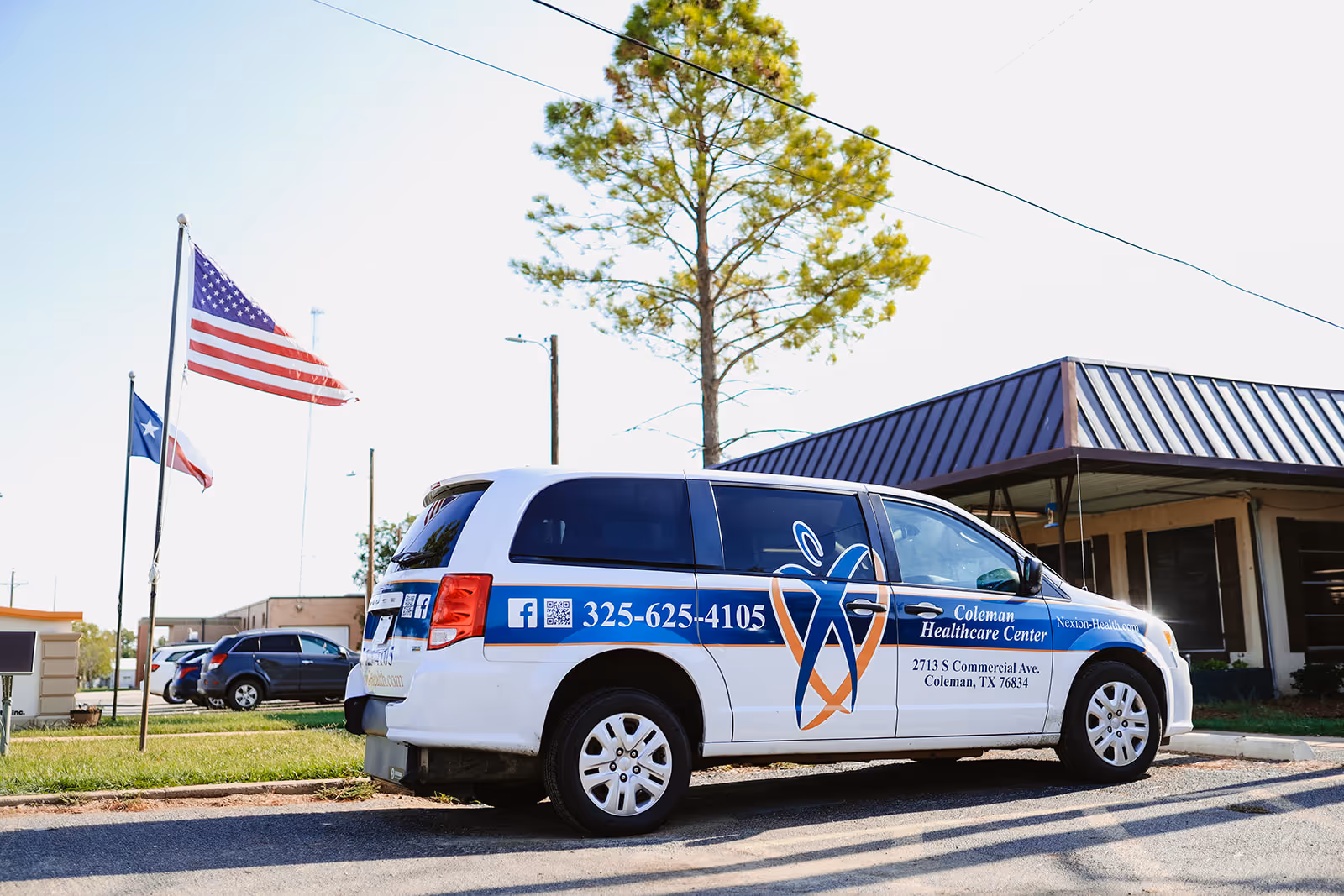 A white van with blue and orange branding for Coleman Healthcare Center is parked on a street near a building with a dark metal roof. Two flagpoles with the American flag and Texas state flag are visible in the background along with a tree and other parked cars.