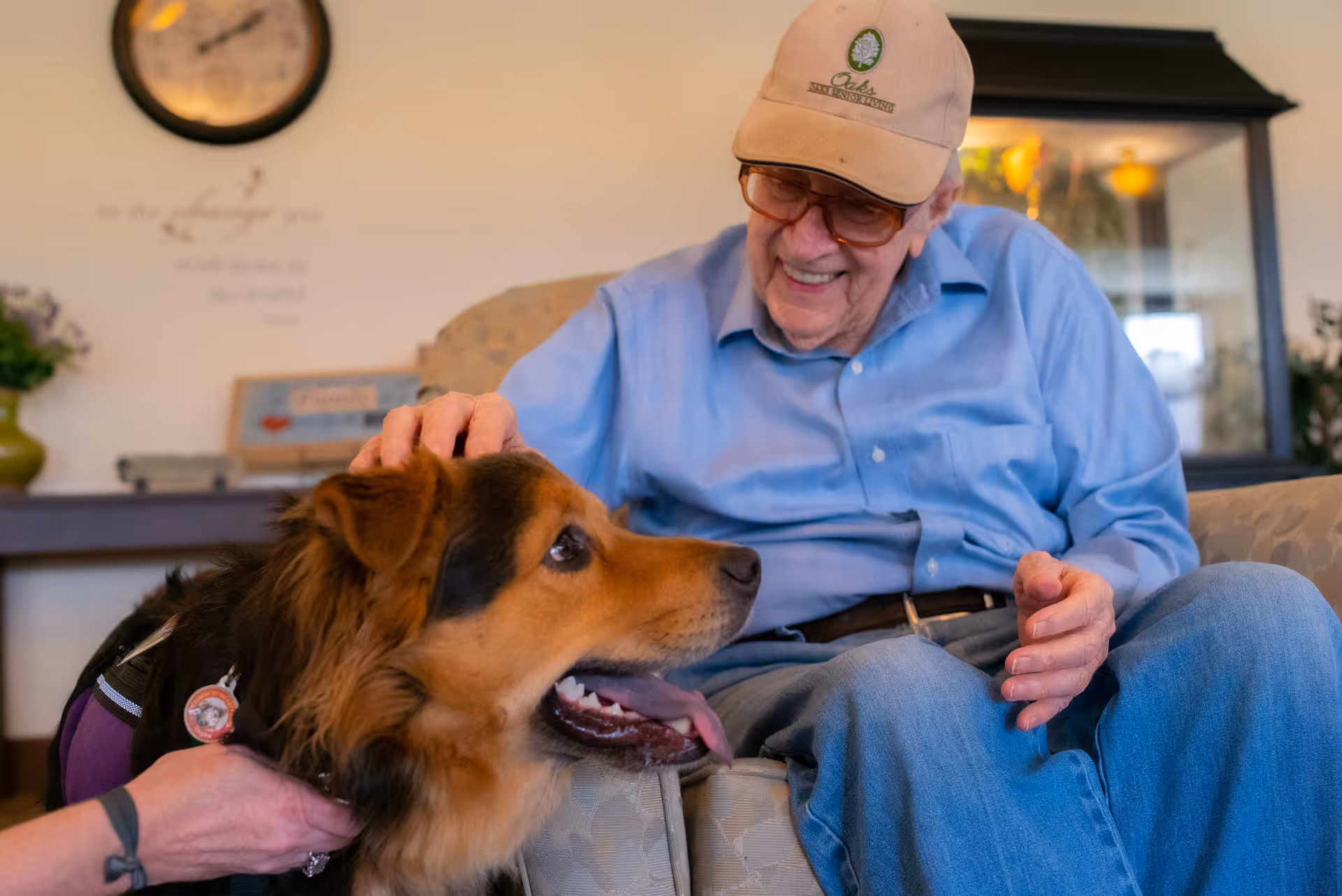 An elderly man wearing a beige Oaks at Post Road cap and blue shirt sits on a couch smiling and petting a large brown and black dog with its tongue out. The setting appears to be a cozy indoor living area with a clock and decorative items in the background.