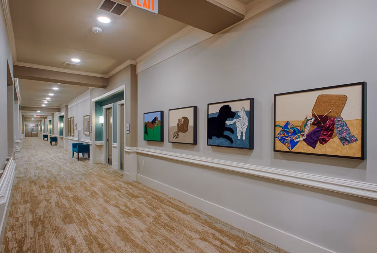 A long, well-lit hallway in an assisted living facility with beige walls and carpeted floor. The right wall features four framed textile art pieces depicting a horse, a basket with yarn, a black dog with a white cat, and a basket with colorful fabric. There are blue chairs along the left side near doorways, and an exit sign is visible on the ceiling.