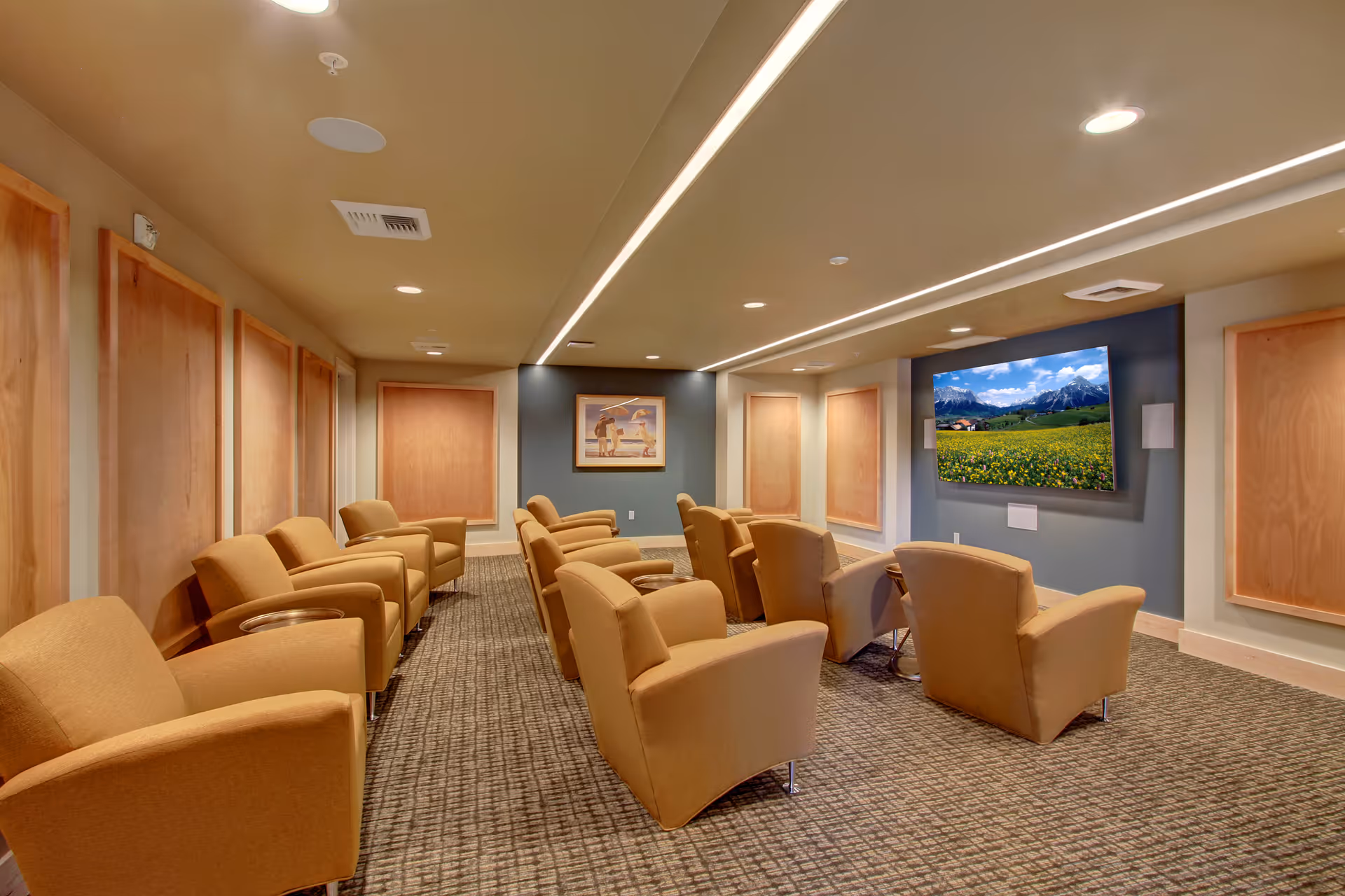A cozy media room with two rows of comfortable tan armchairs facing a large flat-screen TV mounted on a blue accent wall. The room has recessed lighting, carpeted floor, and wooden panel decorations on the walls.
