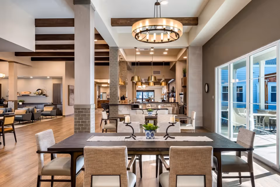 A spacious and modern dining area in a senior living facility with a large dark wooden table surrounded by beige upholstered chairs. The room features wooden ceiling beams, a circular chandelier, and large windows letting in natural light. In the background, there is a kitchen area with pendant lights and additional seating. The space has a warm and inviting atmosphere with light wood flooring and neutral-colored walls.