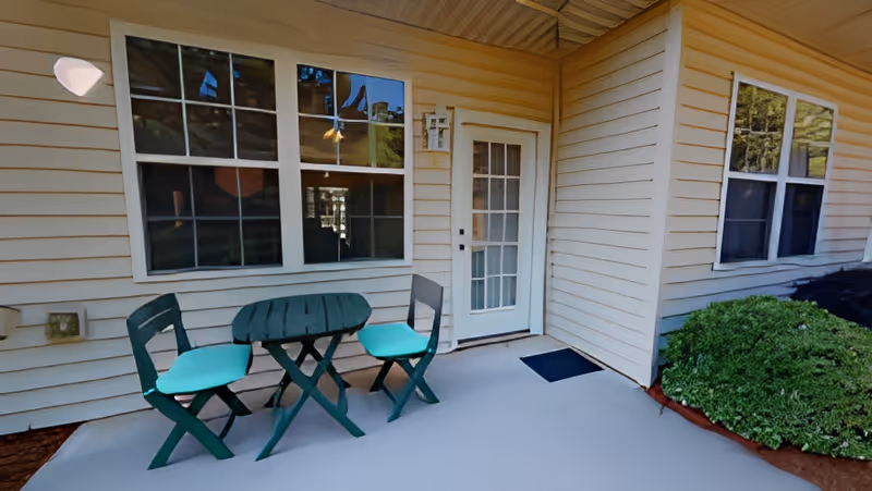 Covered patio outside a unit featuring a small green table with two matching chairs, a glass-paned door, windows, and nearby shrubs.