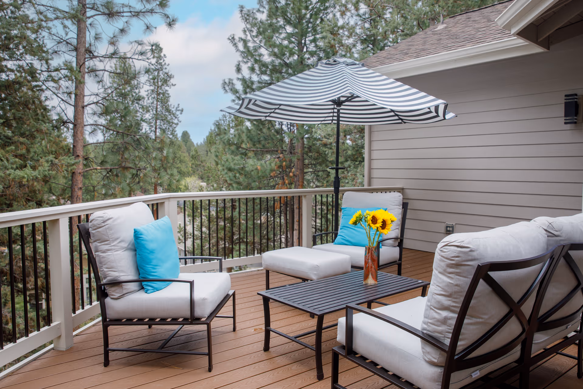 Outdoor patio area with cushioned chairs and a small table holding a vase with sunflowers. A striped umbrella provides shade, and the patio overlooks a forested area with tall pine trees.