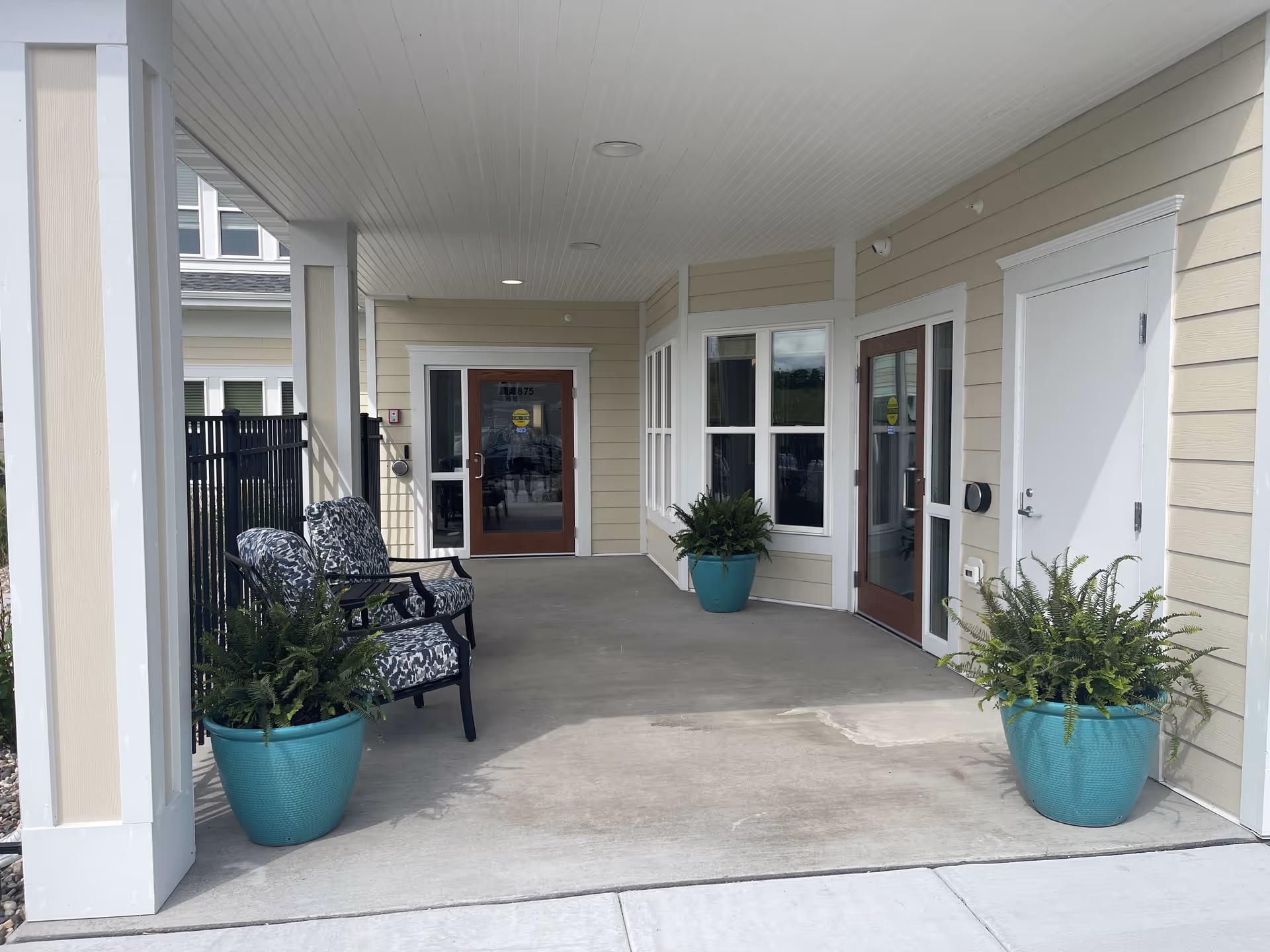 Covered entrance area of a senior living facility with two patterned cushioned chairs and three large teal planters containing green plants. The area has beige siding walls, white trim, and two glass doors with wooden frames. The floor is concrete and the ceiling is white with recessed lighting.