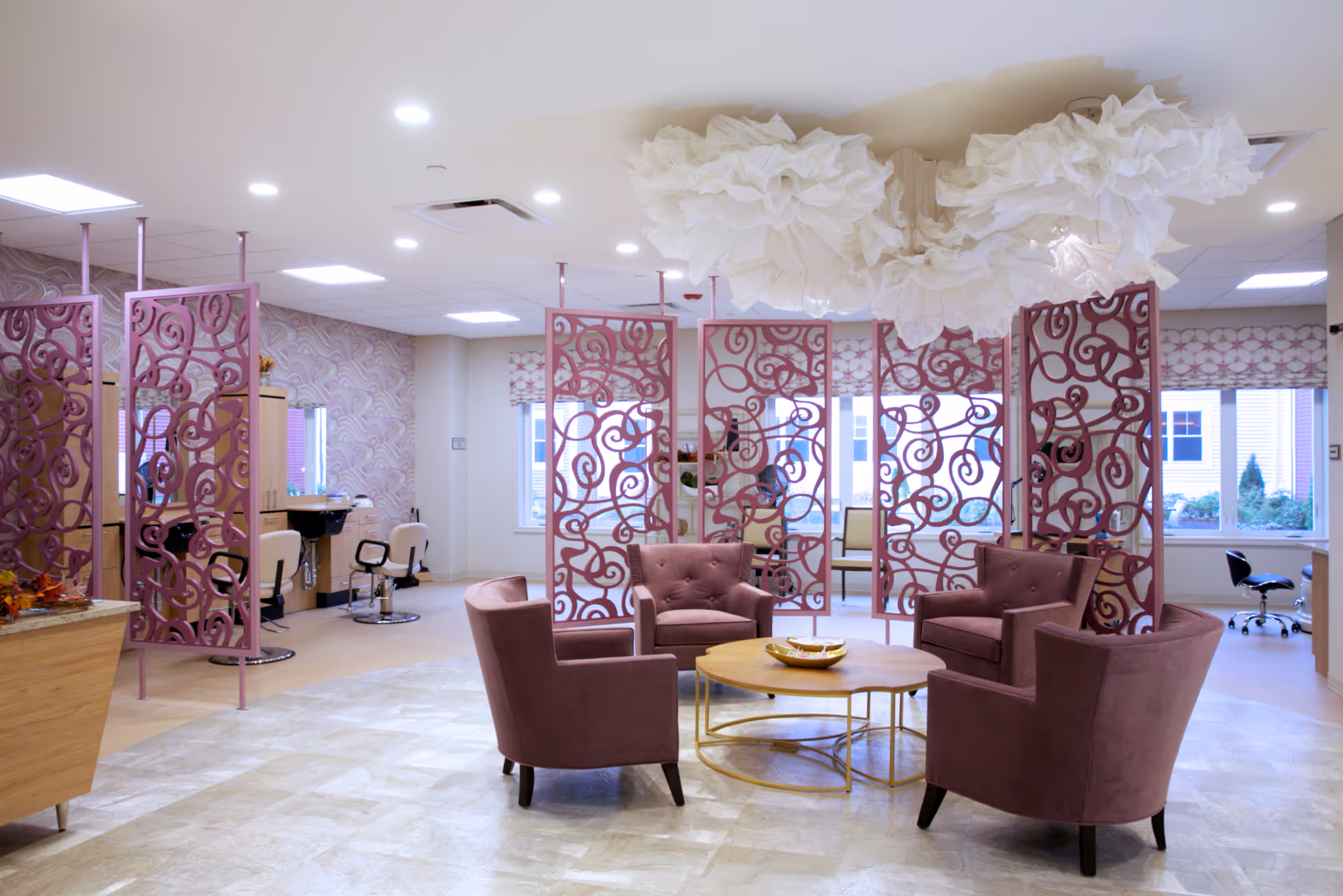 A bright and spacious interior room with four mauve upholstered armchairs arranged around a circular wooden coffee table. Decorative purple metal screens separate the seating area from a salon area with styling chairs and mirrors. Large windows with patterned valances let in natural light, and a unique white cloud-like ceiling decoration hangs above the seating area.