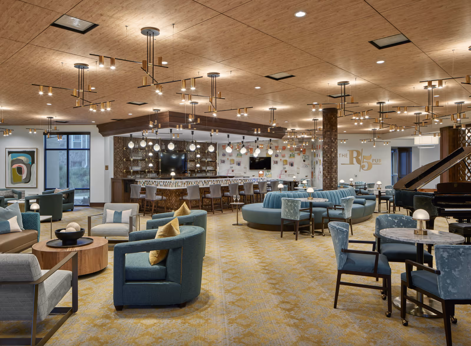 Spacious, well-lit communal lounge and bar area with multiple seating groupings, tables, and a grand piano.