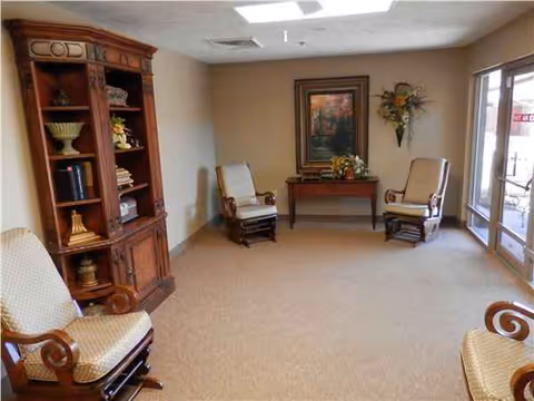 Cozy, well-lit lounge with chairs, a wooden cabinet, floral arrangements, and glass doors.