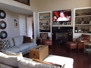 Sunlit living room with sofas and leather armchairs arranged around a central wicker coffee table, a fireplace with a TV above and built-in shelving on either side.