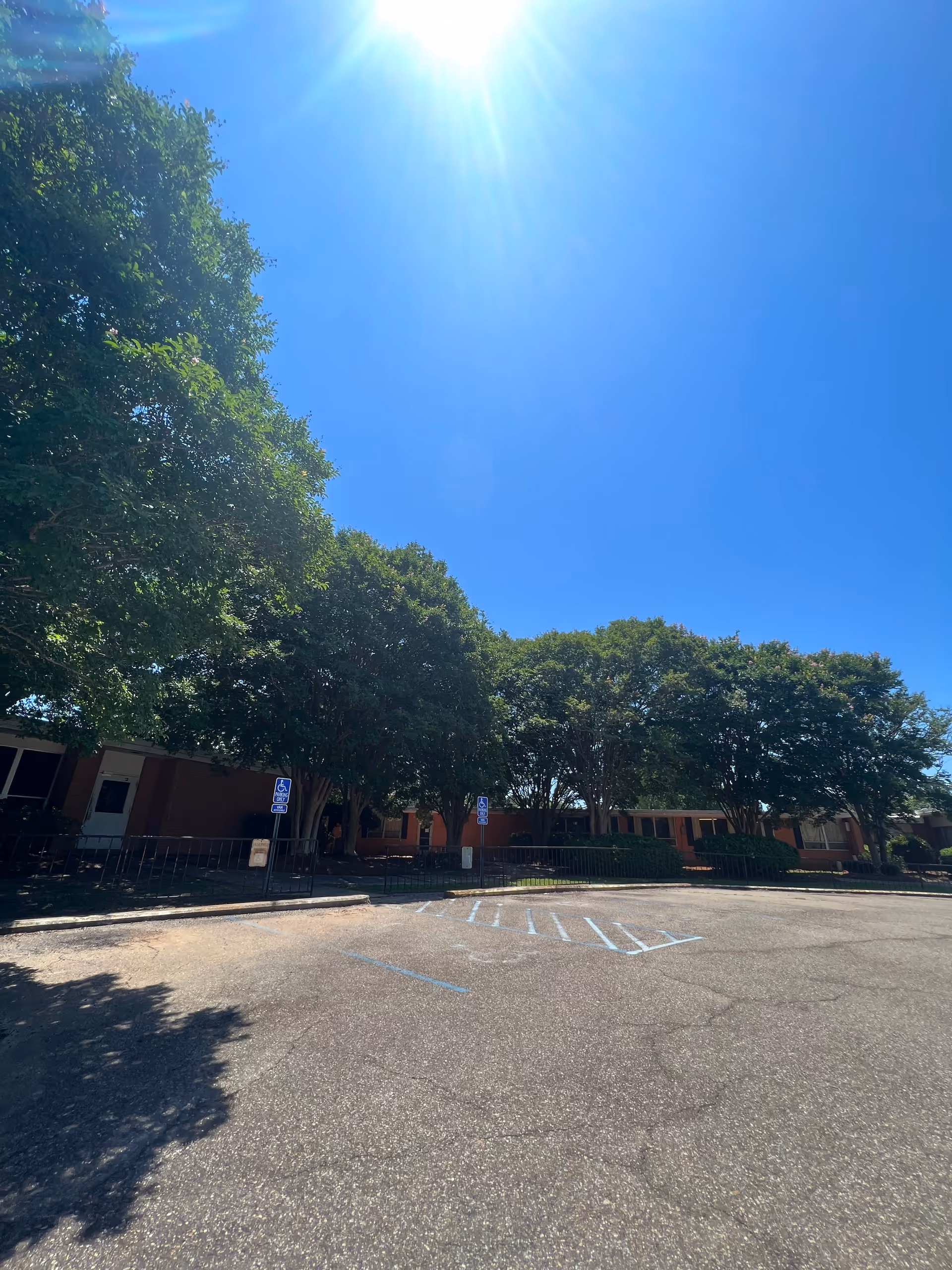 Sunny exterior view of a low brick building partially shaded by trees with an empty parking lot in front.