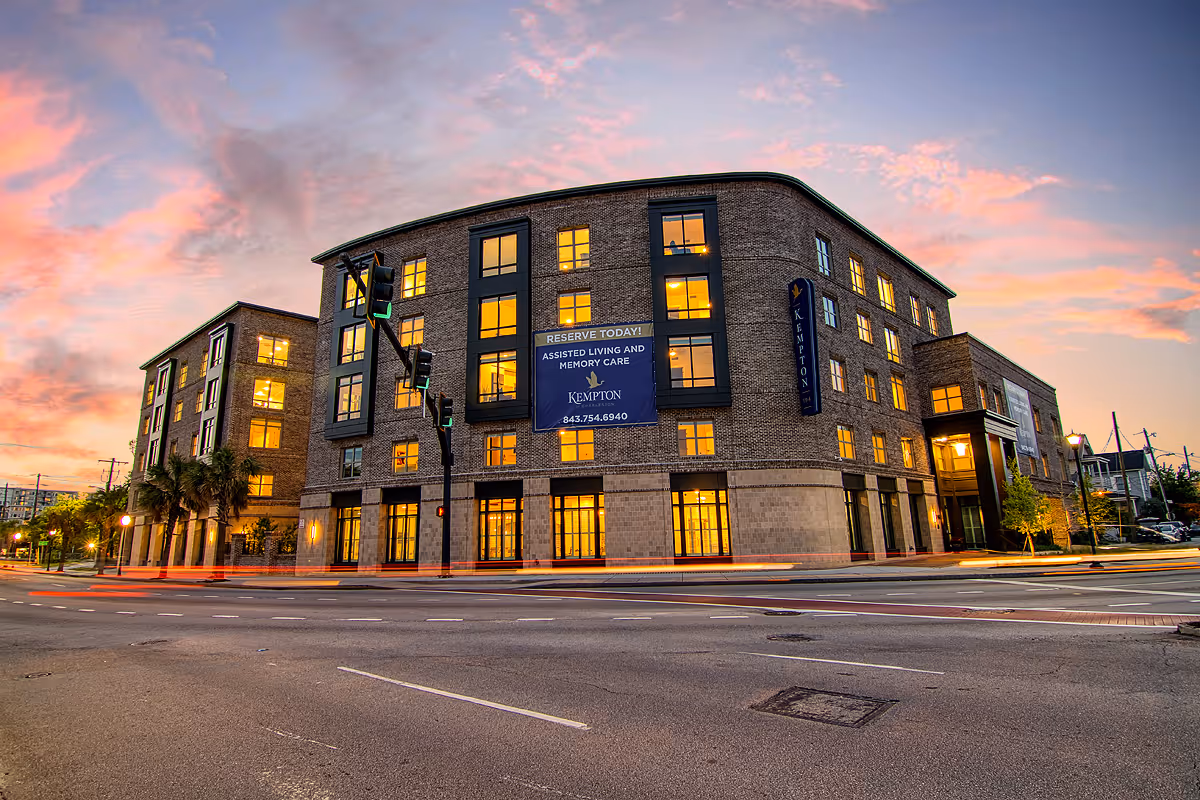 Exterior view of a multi-story brick building at sunset with illuminated windows and a large sign advertising assisted living and memory care at Kempton of Charleston. The building is located at a street intersection with traffic lights and some light trails from passing vehicles.