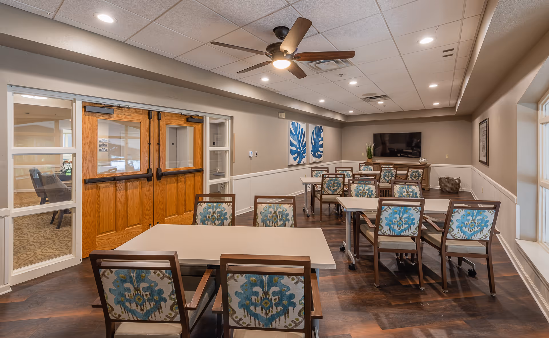 A well-lit room with several tables and chairs arranged in rows. The chairs have a floral patterned upholstery. There is a ceiling fan with a light in the center of the ceiling. On one wall, there are two blue leaf-themed artworks and a flat-screen TV mounted above a wooden cabinet. The room has wooden double doors with glass panels and windows on either side, allowing a view into an adjacent room.