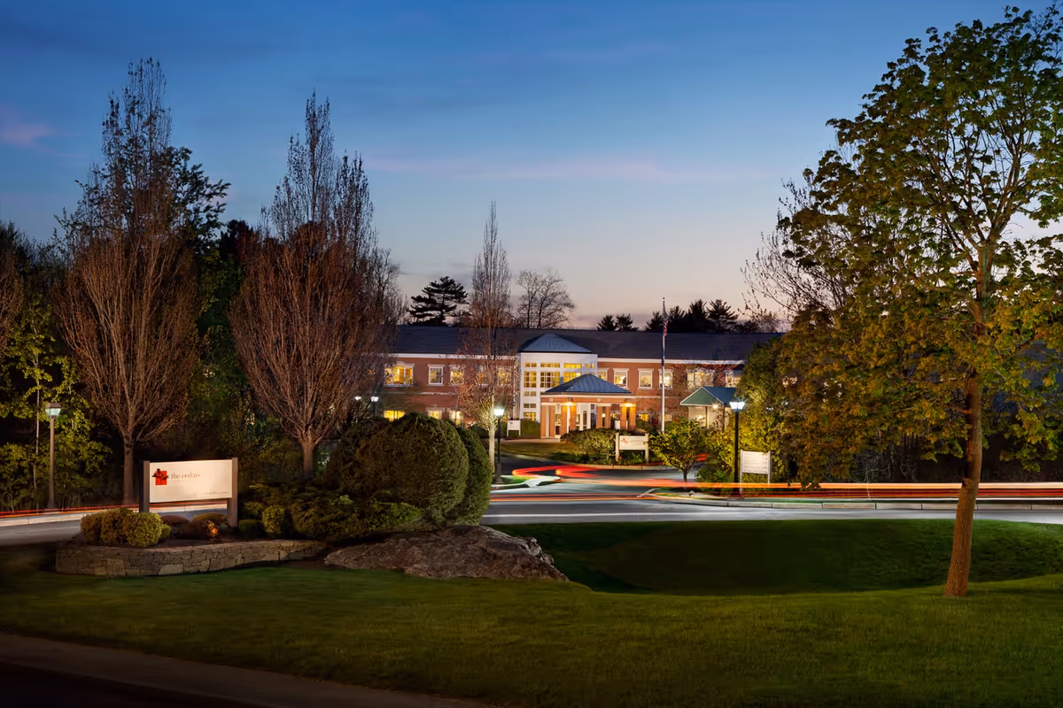 Front exterior of a brick senior living facility at dusk with a landscaped entrance, illuminated windows, and light trails from passing cars.